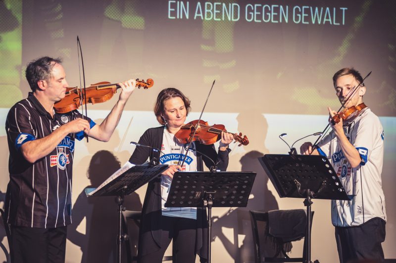 Drei Geigenspieler musizieren auf der Bühne und tragen SK Strum Shirts, zu sehen ist der Slogan "Abend gegen Gewalt" im Hintegrund.